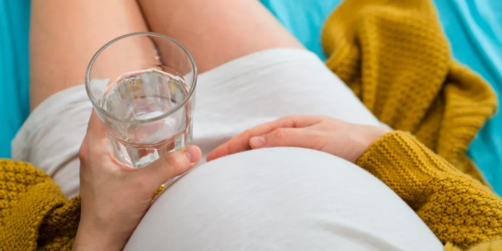 A pregnant woman in a white dress rests on a bed, holding a glass of water. She's relaxed, with a yellow knit blanket beside her on blue sheets.